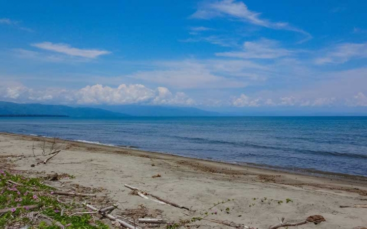 In the foreground, a sandy beach appears in front of a very blue ocean. In the distances, there appears to be mountains.